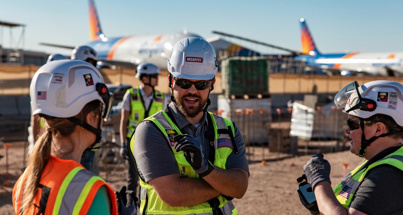 Three people having a discussion at an airport jobsite with planes in the background